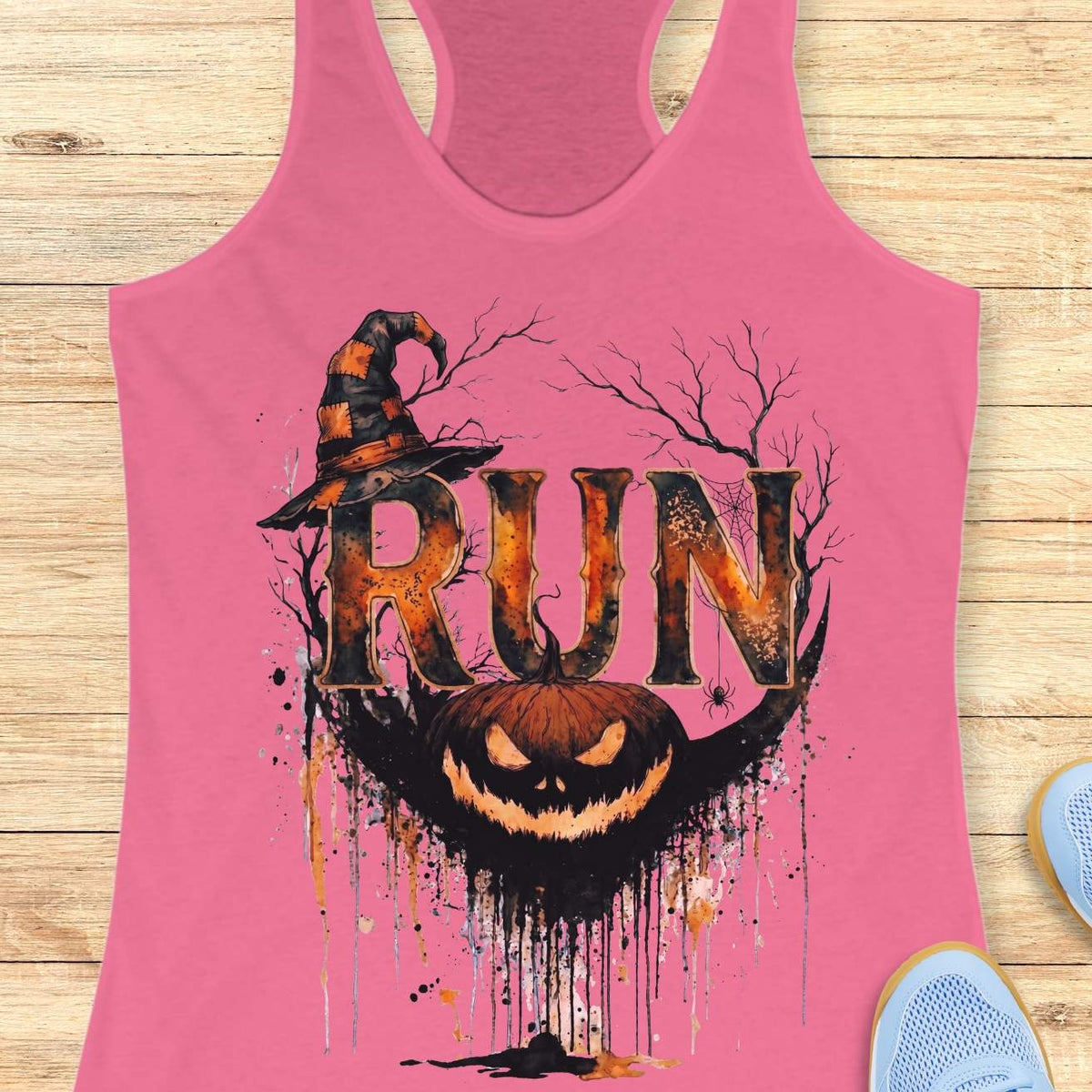 pink racerback tank top featuring a Halloween-themed running design. The graphic displays the word “RUN” in large distressed orange and black letters, topped with a weathered witch hat. Behind the text are bare tree branches, a spider web, and a hanging spider. A grinning jack-o'-lantern with dripping black and orange paint effects sits below the lettering, creating a spooky autumn aesthetic. The tank top is displayed on a light wooden background with partial blue running shoes visible in the corners.