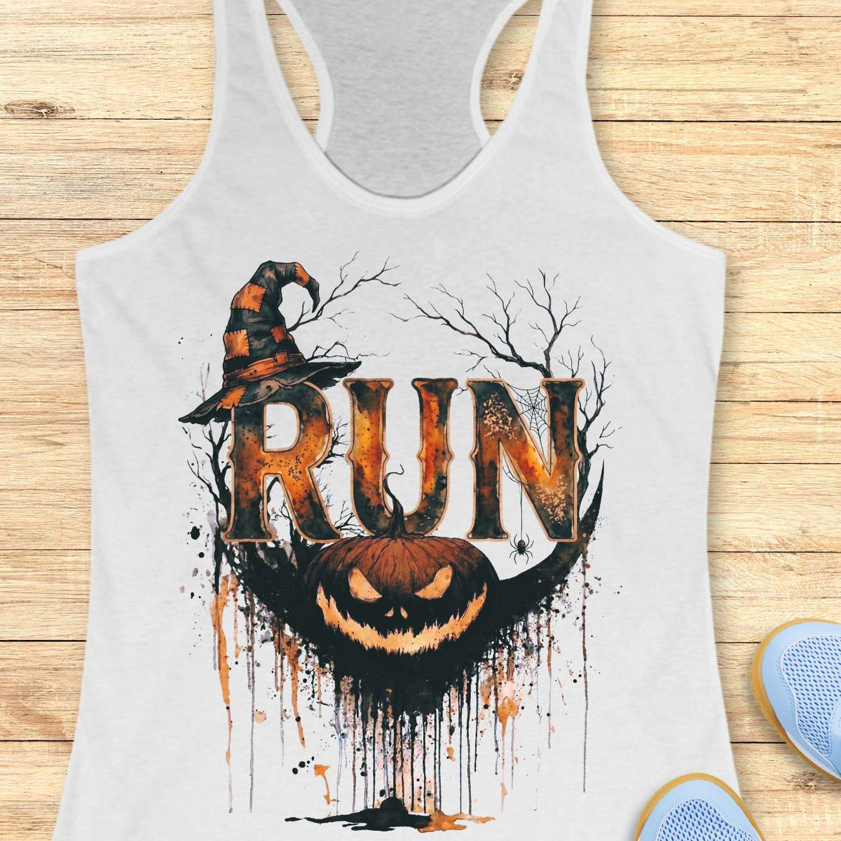 white racerback tank top featuring a Halloween-themed running design. The graphic displays the word “RUN” in large distressed orange and black letters, topped with a weathered witch hat. Behind the text are bare tree branches, a spider web, and a hanging spider. A grinning jack-o'-lantern with dripping black and orange paint effects sits below the lettering, creating a spooky autumn aesthetic. The tank top is displayed on a light wooden background with partial blue running shoes visible in the corners.