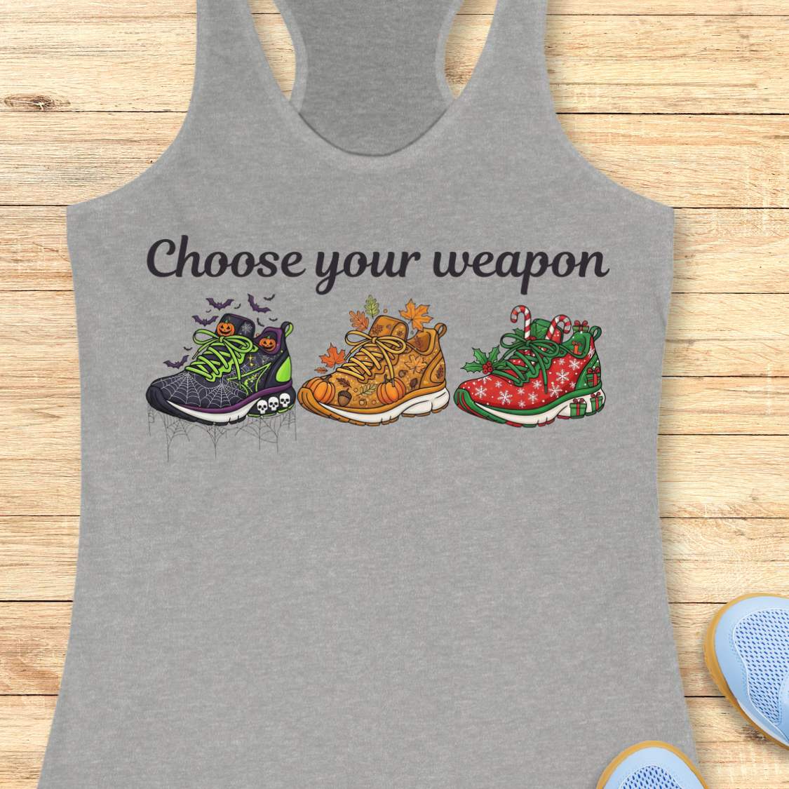 Heather gray women's racerback tank top displayed on a light wood background. The design features the phrase “Choose your weapon” in black script lettering above three themed running shoes. The first shoe has a Halloween design with spiderwebs, pumpkins, bats, and skulls. The second shoe has an autumn theme with pumpkins, leaves, and acorns. The third shoe has a Christmas theme with candy canes, holly, snowflakes, and gift boxes. Partial running shoes are visible in the lower corners of the image.