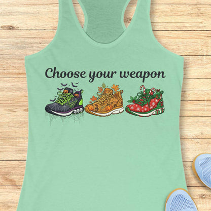 mint women's racerback tank top displayed on a light wood background. The design features the phrase “Choose your weapon” in black script lettering above three themed running shoes. The first shoe has a Halloween design with spiderwebs, pumpkins, bats, and skulls. The second shoe has an autumn theme with pumpkins, leaves, and acorns. The third shoe has a Christmas theme with candy canes, holly, snowflakes, and gift boxes. Partial running shoes are visible in the lower corners of the image.