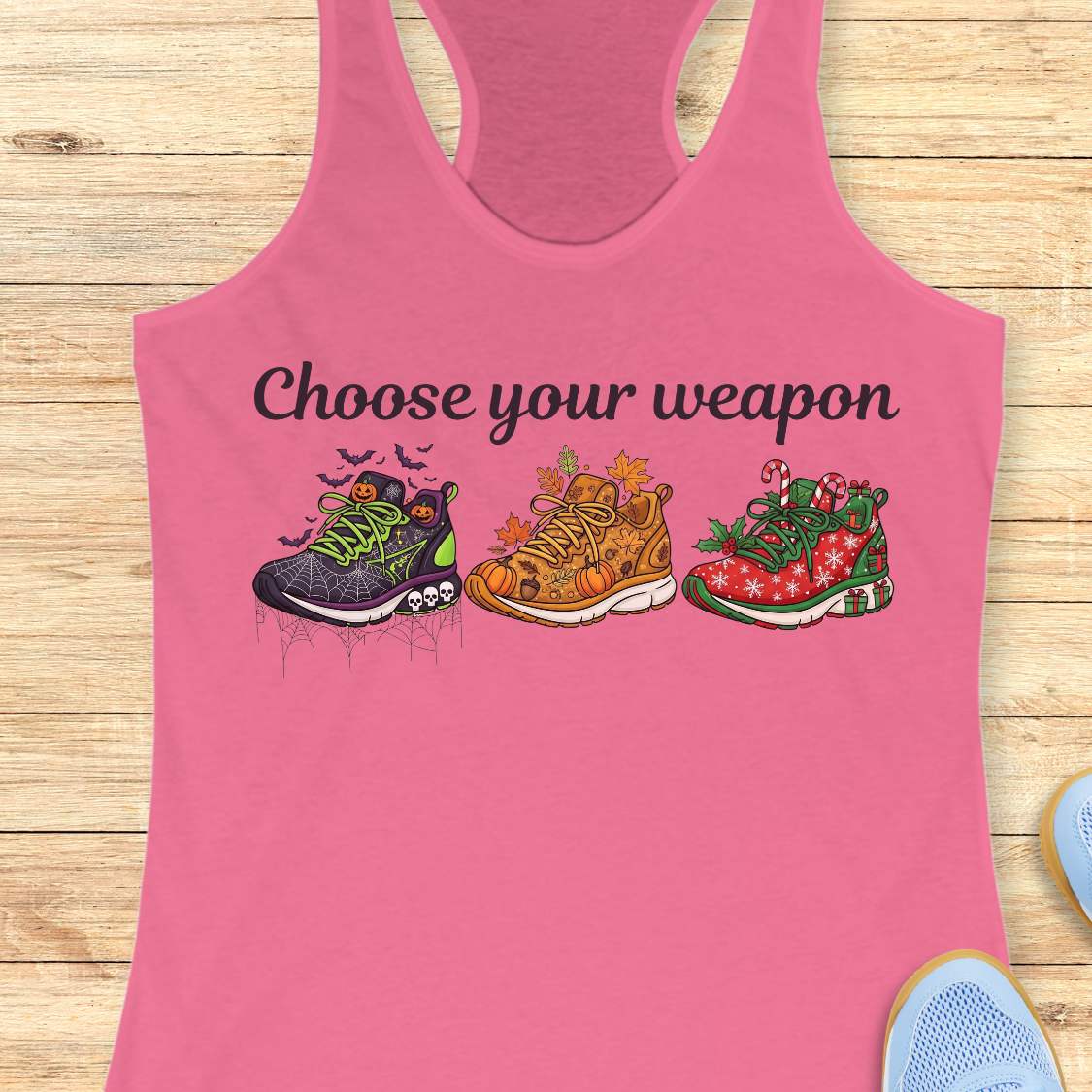 pink women's racerback tank top displayed on a light wood background. The design features the phrase “Choose your weapon” in black script lettering above three themed running shoes. The first shoe has a Halloween design with spiderwebs, pumpkins, bats, and skulls. The second shoe has an autumn theme with pumpkins, leaves, and acorns. The third shoe has a Christmas theme with candy canes, holly, snowflakes, and gift boxes. Partial running shoes are visible in the lower corners of the image.