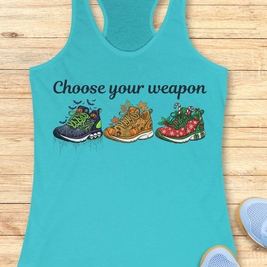 blue women's racerback tank top displayed on a light wood background. The design features the phrase “Choose your weapon” in black script lettering above three themed running shoes. The first shoe has a Halloween design with spiderwebs, pumpkins, bats, and skulls. The second shoe has an autumn theme with pumpkins, leaves, and acorns. The third shoe has a Christmas theme with candy canes, holly, snowflakes, and gift boxes. Partial running shoes are visible in the lower corners of the image.