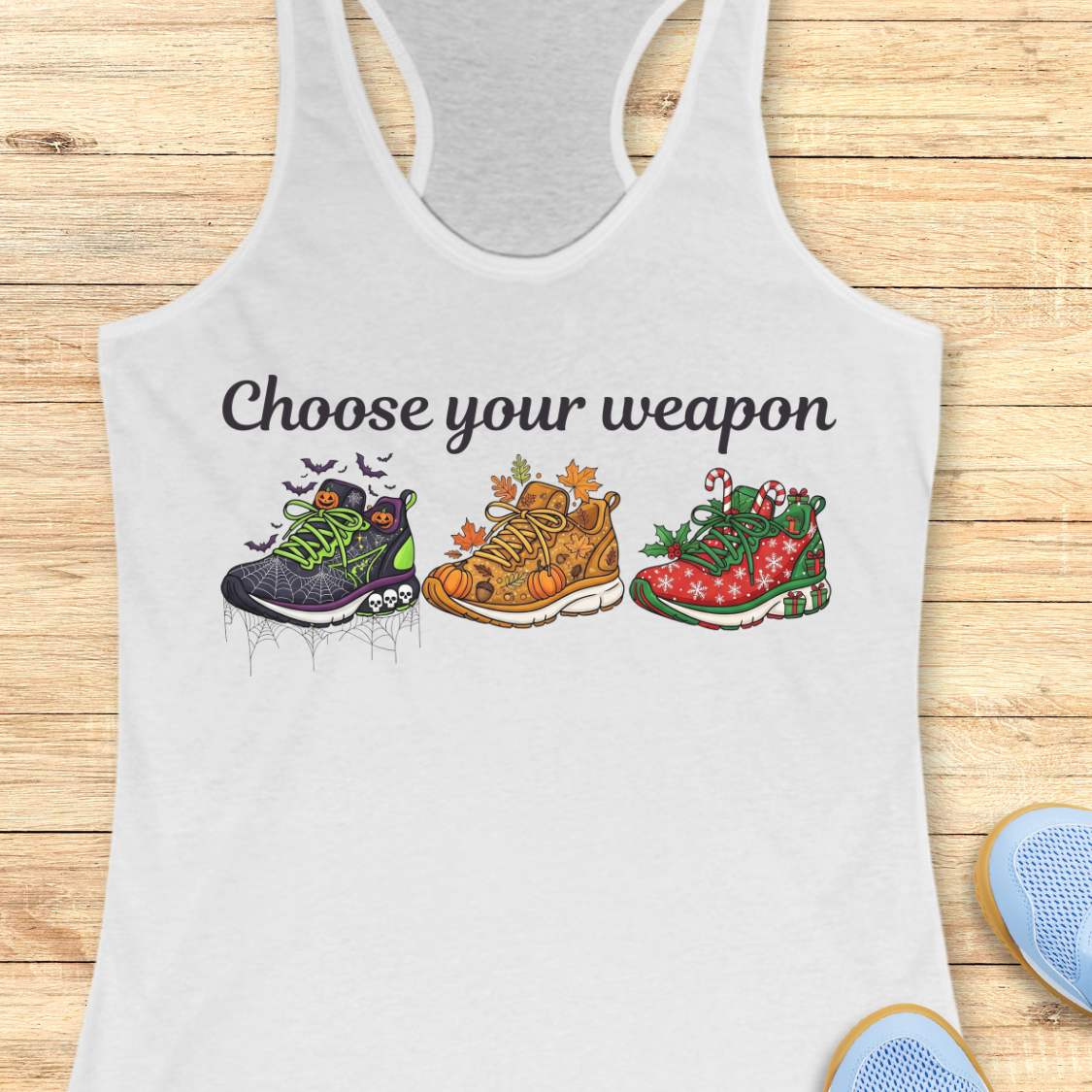 white women's racerback tank top displayed on a light wood background. The design features the phrase “Choose your weapon” in black script lettering above three themed running shoes. The first shoe has a Halloween design with spiderwebs, pumpkins, bats, and skulls. The second shoe has an autumn theme with pumpkins, leaves, and acorns. The third shoe has a Christmas theme with candy canes, holly, snowflakes, and gift boxes. Partial running shoes are visible in the lower corners of the image.