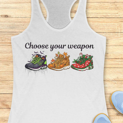white women's racerback tank top displayed on a light wood background. The design features the phrase “Choose your weapon” in black script lettering above three themed running shoes. The first shoe has a Halloween design with spiderwebs, pumpkins, bats, and skulls. The second shoe has an autumn theme with pumpkins, leaves, and acorns. The third shoe has a Christmas theme with candy canes, holly, snowflakes, and gift boxes. Partial running shoes are visible in the lower corners of the image.