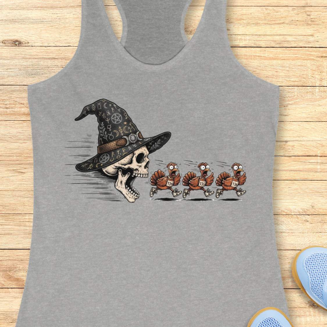 A grey racerback tank top laid flat on a light wood surface, featuring a graphic print of a skull wearing a witch hat with three cartoon turkeys chasing it. Partial shoes are visible at the bottom right as styling props.