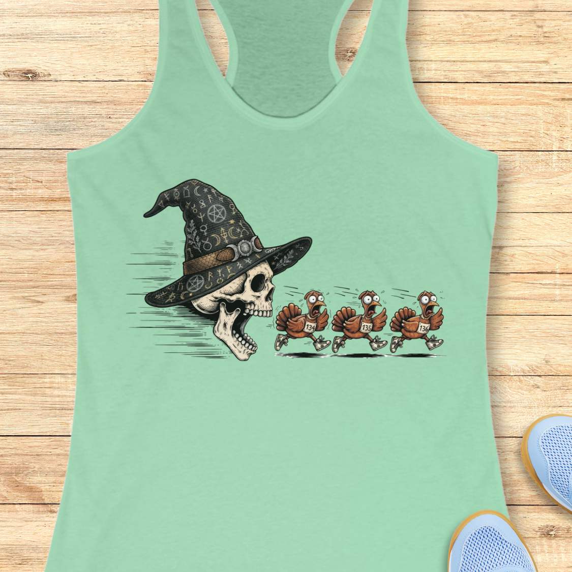 A mint racerback tank top laid flat on a light wood surface, featuring a graphic print of a skull wearing a witch hat with three cartoon turkeys chasing it. Partial shoes are visible at the bottom right as styling props.