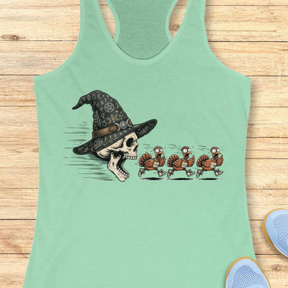 A mint racerback tank top laid flat on a light wood surface, featuring a graphic print of a skull wearing a witch hat with three cartoon turkeys chasing it. Partial shoes are visible at the bottom right as styling props.