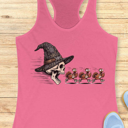 A pink racerback tank top laid flat on a light wood surface, featuring a graphic print of a skull wearing a witch hat with three cartoon turkeys chasing it. Partial shoes are visible at the bottom right as styling props.