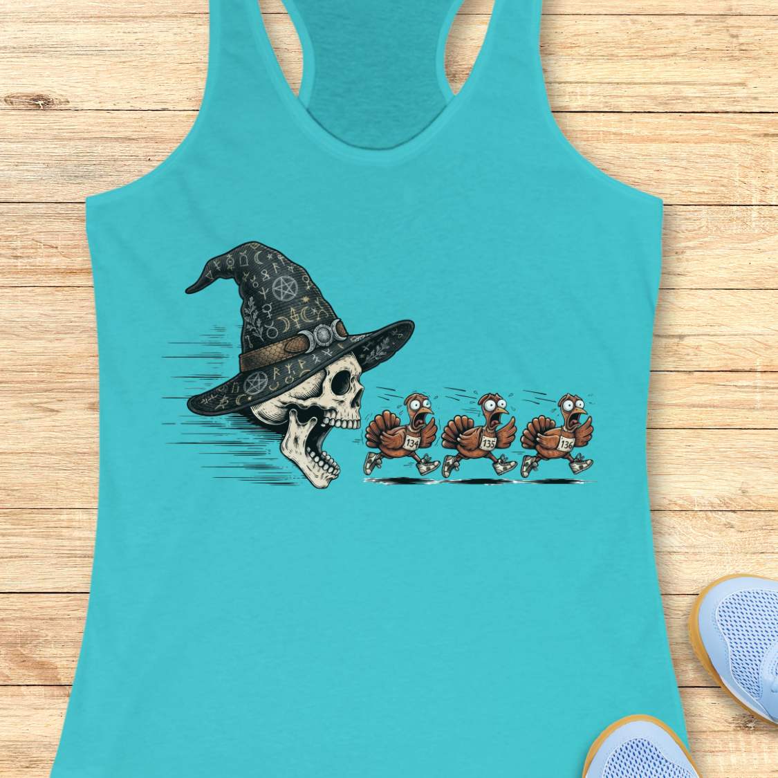 A turquoise racerback tank top laid flat on a light wood surface, featuring a graphic print of a skull wearing a witch hat with three cartoon turkeys chasing it. Partial shoes are visible at the bottom right as styling props.
