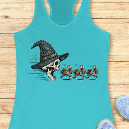 A turquoise racerback tank top laid flat on a light wood surface, featuring a graphic print of a skull wearing a witch hat with three cartoon turkeys chasing it. Partial shoes are visible at the bottom right as styling props.