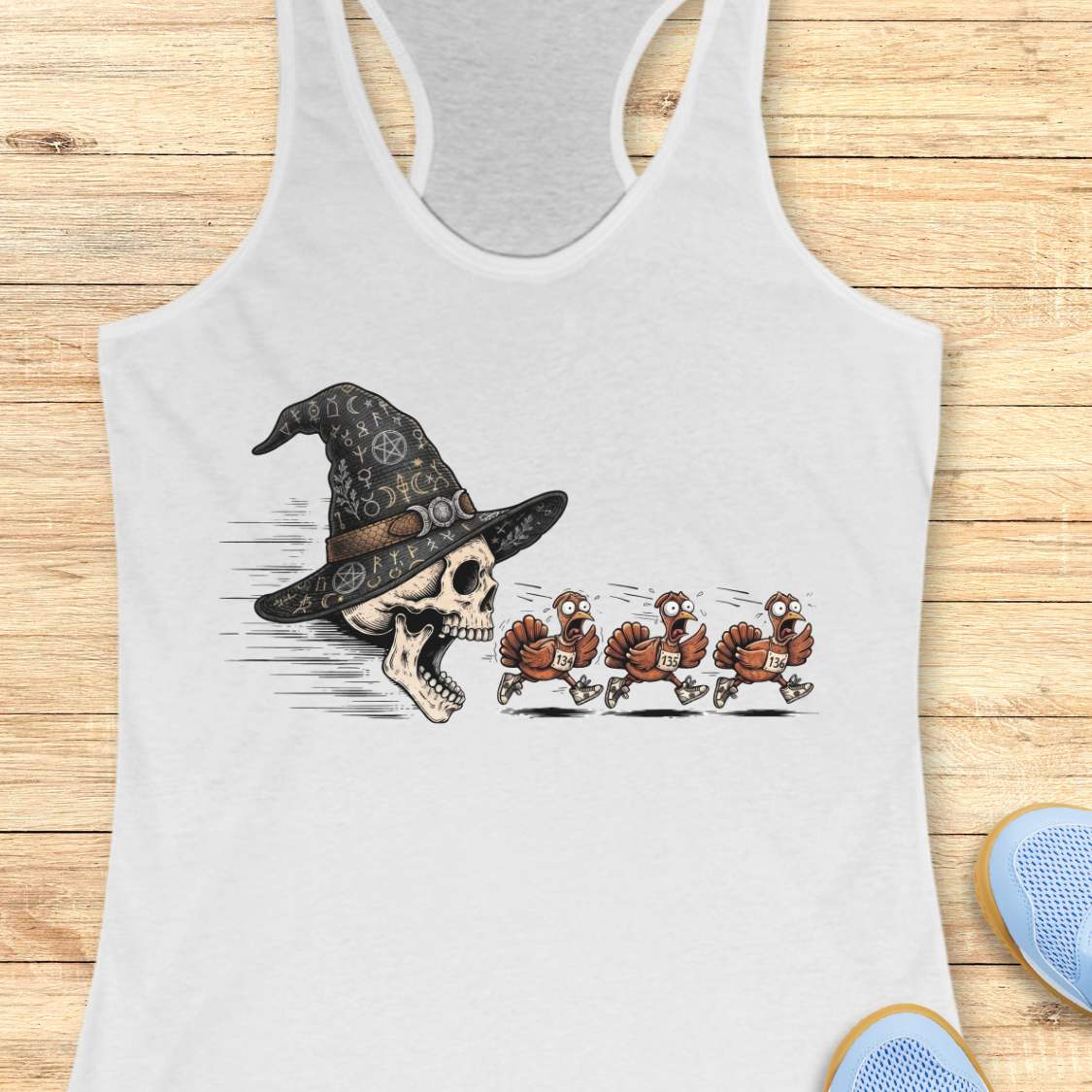 A white racerback tank top laid flat on a light wood surface, featuring a graphic print of a skull wearing a witch hat with three cartoon turkeys chasing it. Partial shoes are visible at the bottom right as styling props.