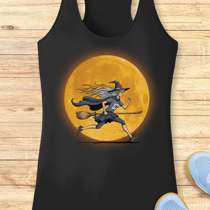 A black racerback tank top laid flat on a light wood surface, featuring a large graphic of a witch running on a broomstick in front of a glowing orange full moon. Part of a blue shoe is visible at the bottom right.