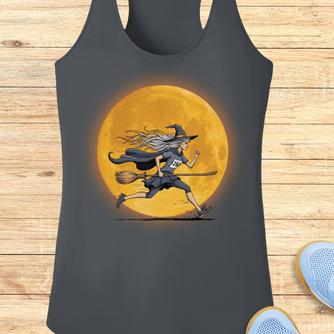 A dark gray racerback tank top featuring a graphic of a witch running on a broomstick in front of a large glowing orange moon, displayed flat on a light wood plank background.