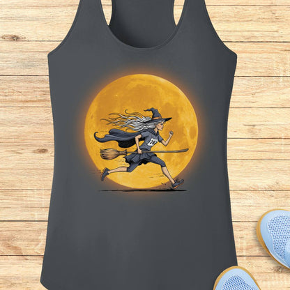 A dark gray racerback tank top featuring a graphic of a witch running on a broomstick in front of a large glowing orange moon, displayed flat on a light wood plank background.
