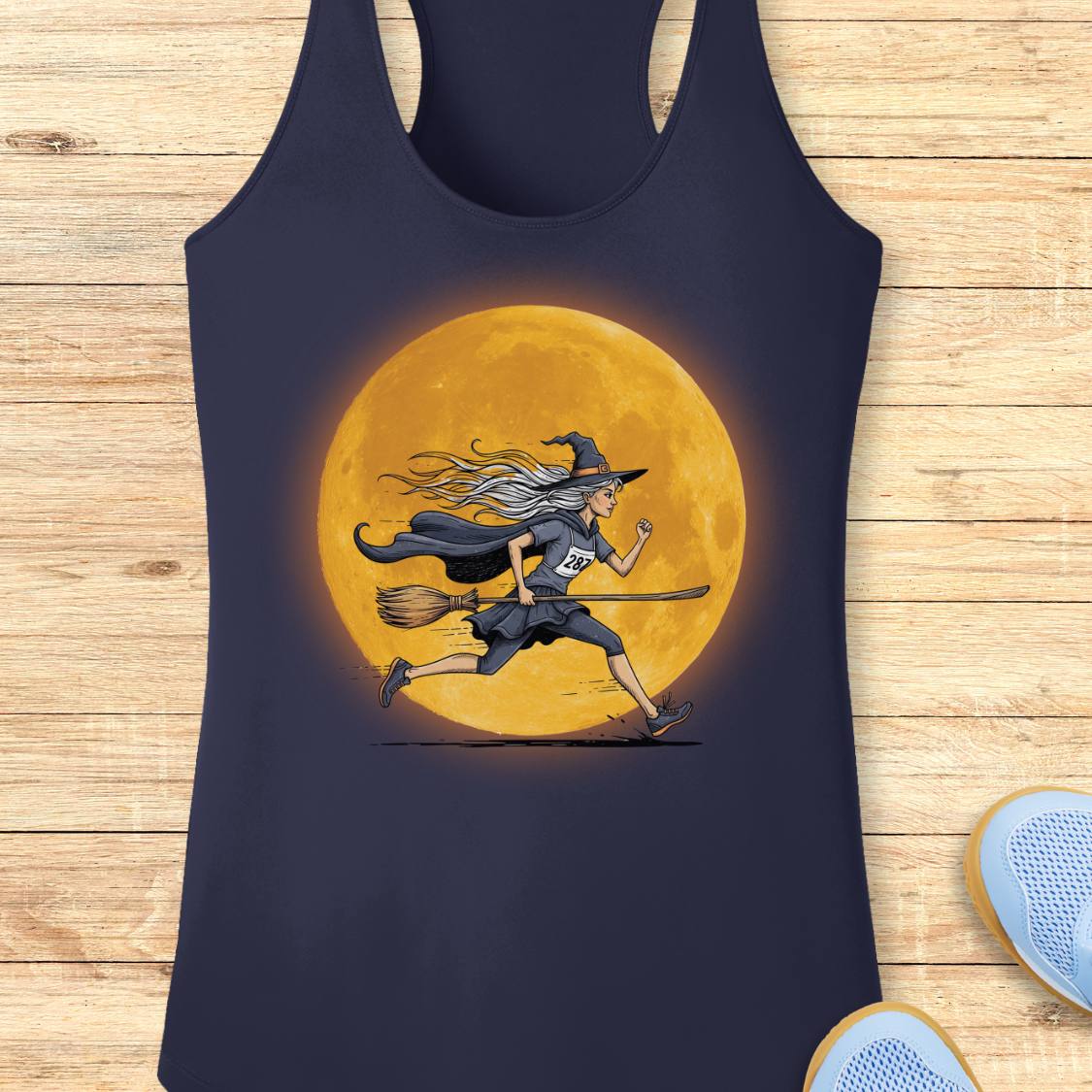 A navy blue racerback tank top featuring a graphic of a witch running on a broomstick in front of a large glowing orange moon, laid flat on a light wood plank background.