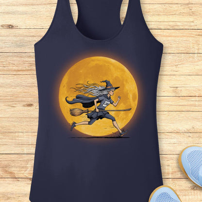 A navy blue racerback tank top featuring a graphic of a witch running on a broomstick in front of a large glowing orange moon, laid flat on a light wood plank background.