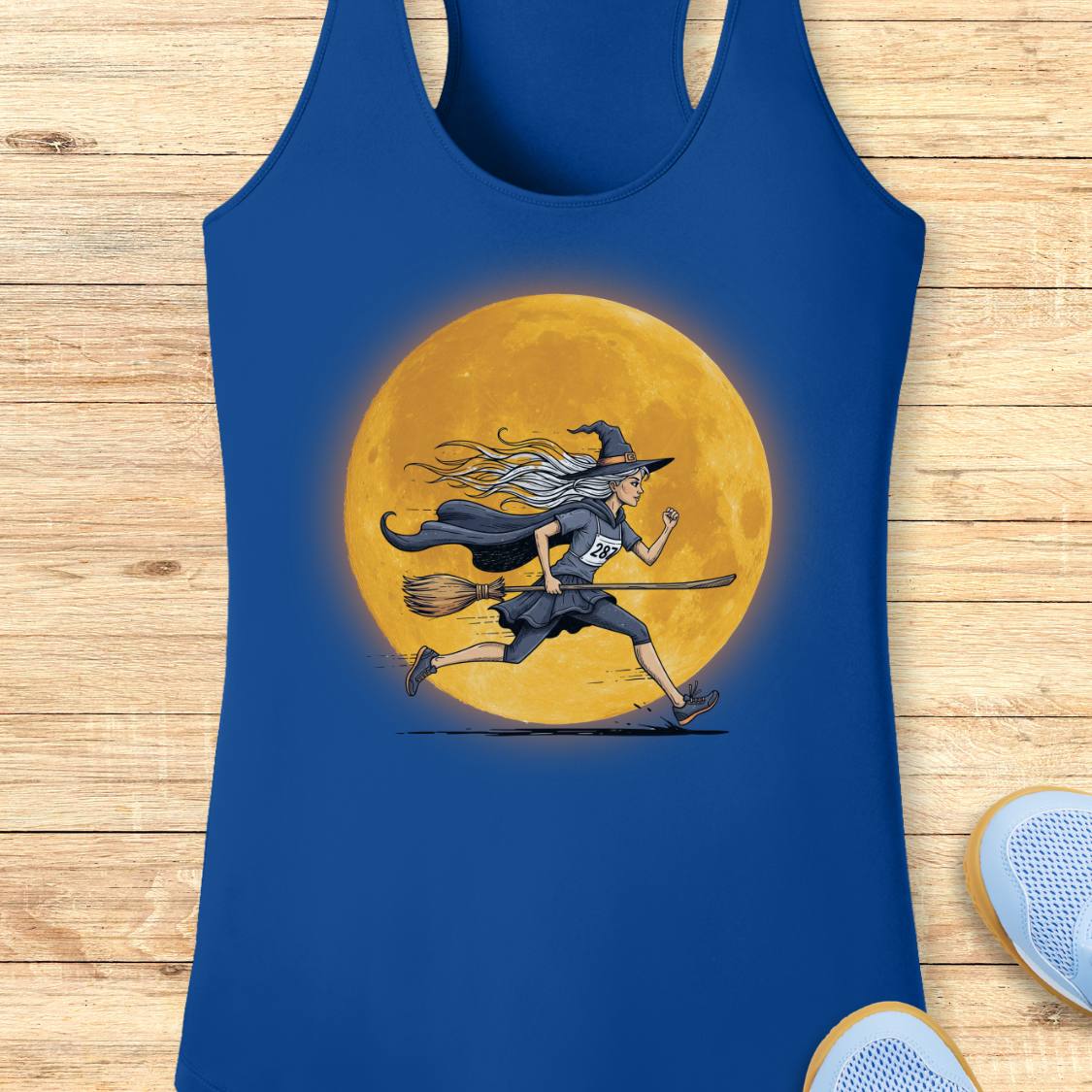 A royal blue racerback tank top featuring a graphic of a witch running on a broomstick in front of a large glowing yellow moon, displayed flat on a light wood plank background.