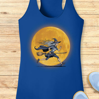 A royal blue racerback tank top featuring a graphic of a witch running on a broomstick in front of a large glowing yellow moon, displayed flat on a light wood plank background.