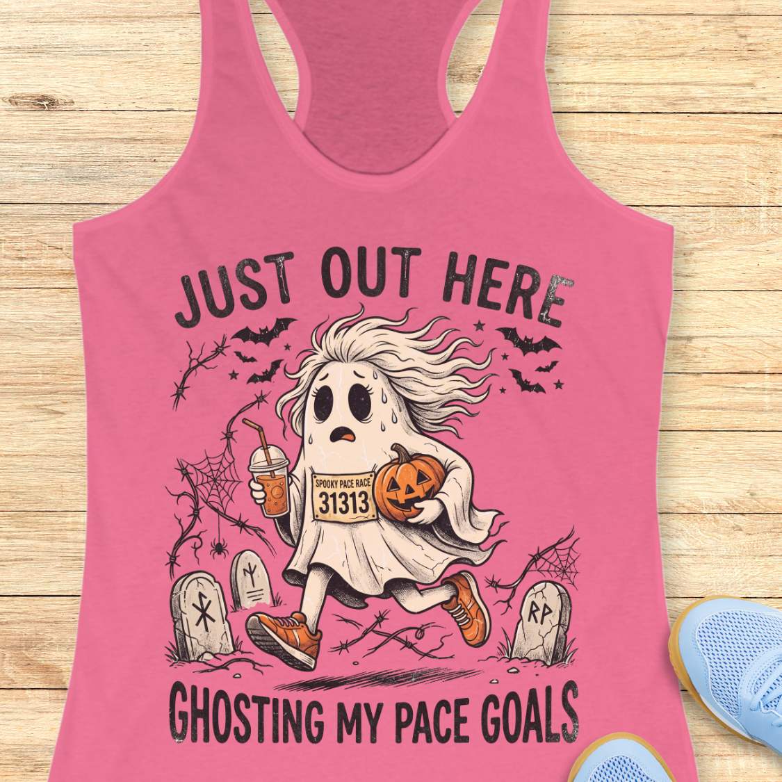 pink Halloween tank top with an image of a running ghost and text 'Just out here ghosting my pace goals', on a wooden surface