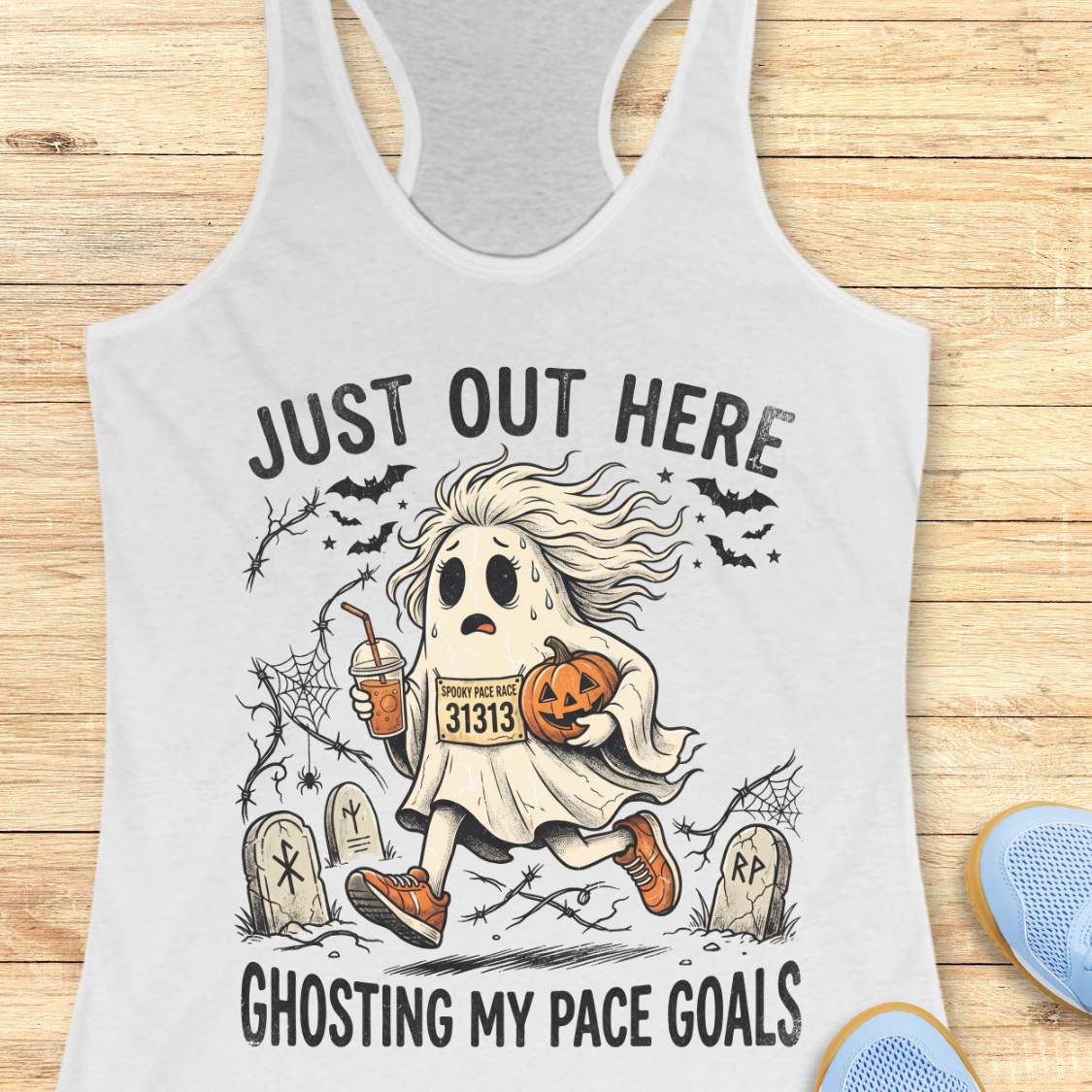 white Halloween tank top with an image of a running ghost and text 'Just out here ghosting my pace goals', on a wooden surface
