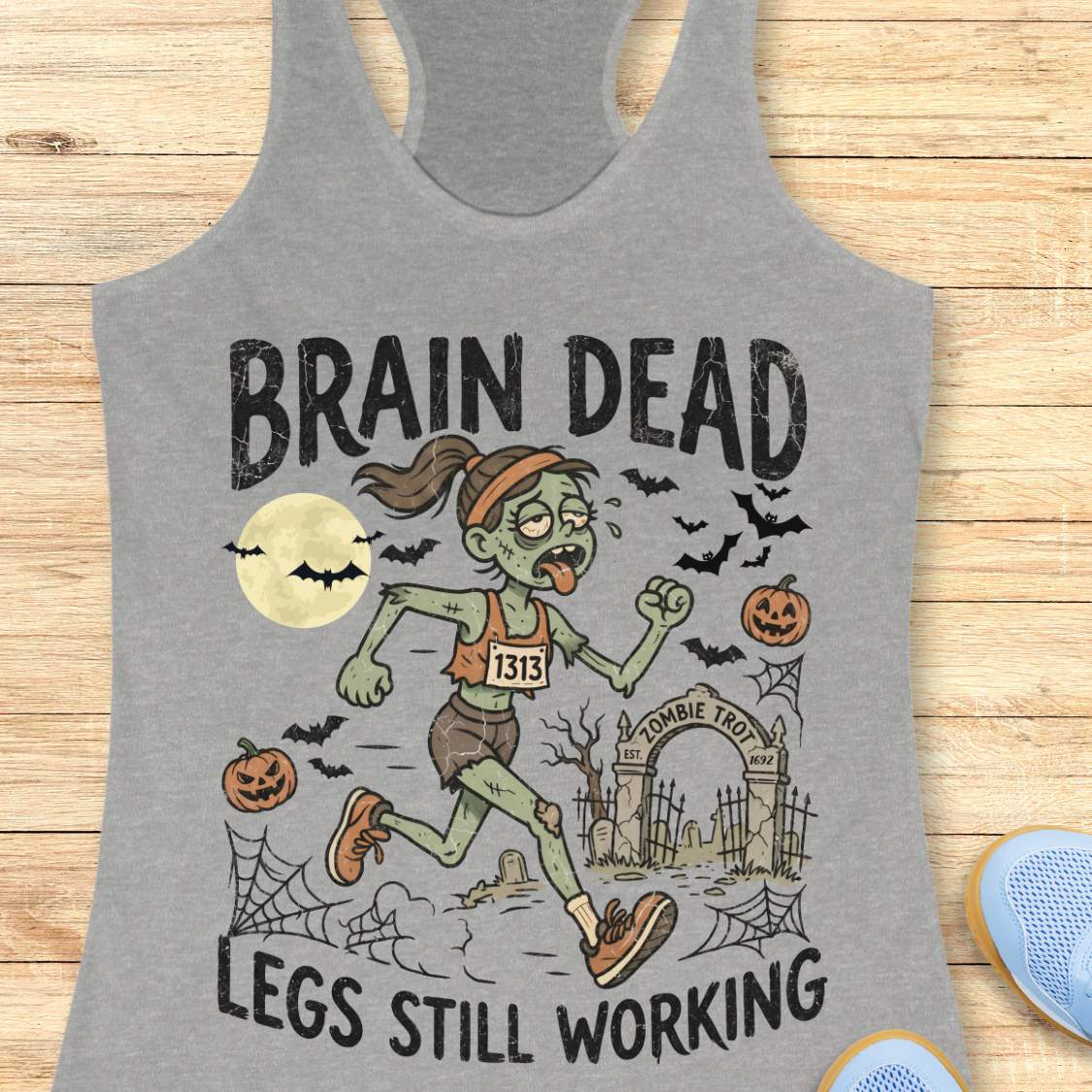 grey Halloween tank top with an image of a running zombie and text 'Brain dead legs still working', on a wooden surface