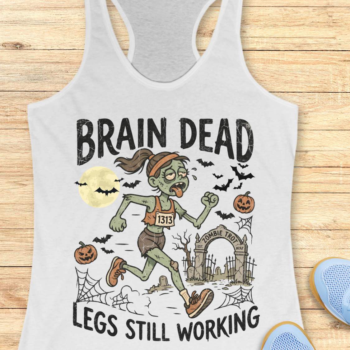 white Halloween tank top with an image of a running zombie and text 'Brain dead legs still working', on a wooden surface