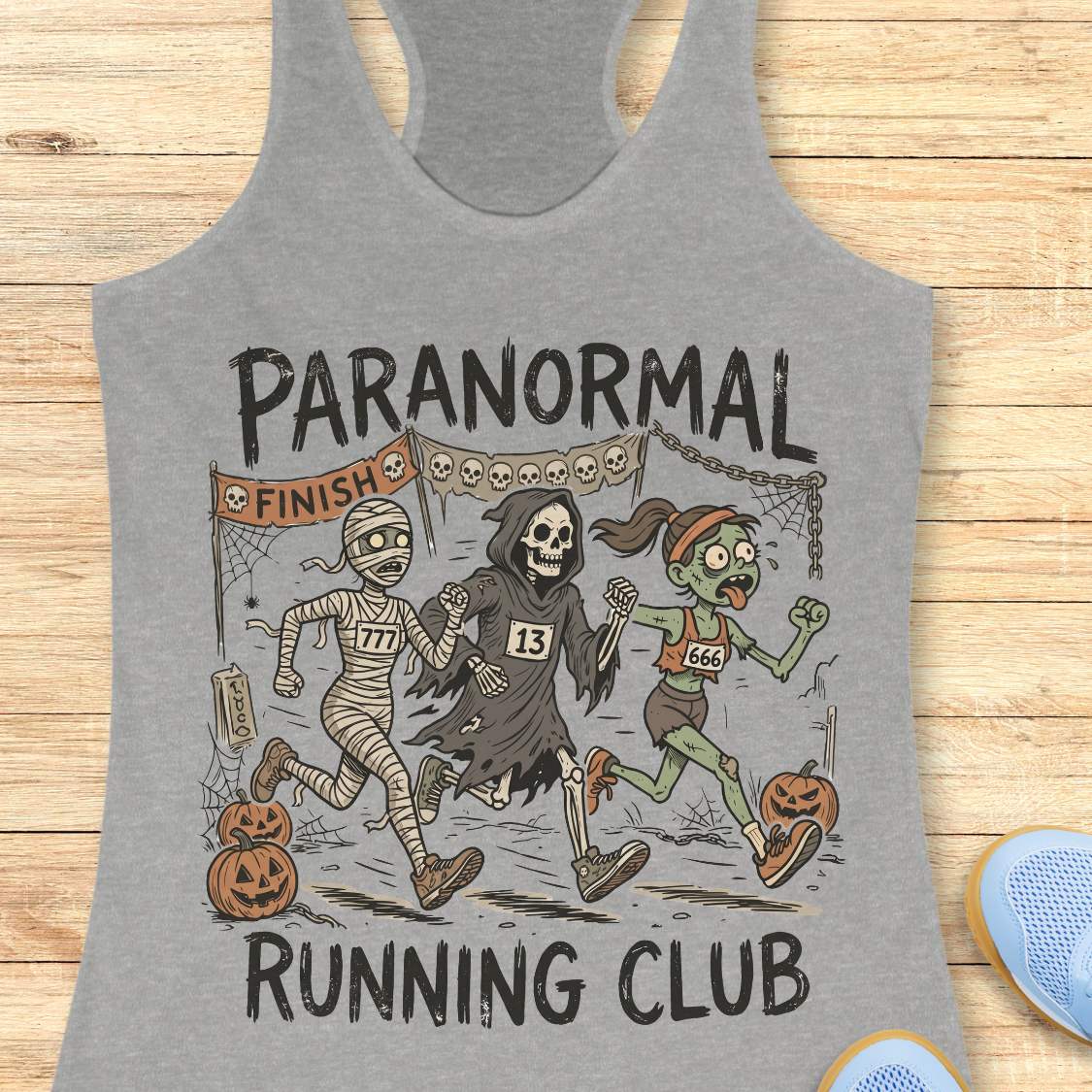 grey Halloween tank top with an image of a running zombie, mummy and reaper,  and text 'Paranormal running club', on a wooden surface