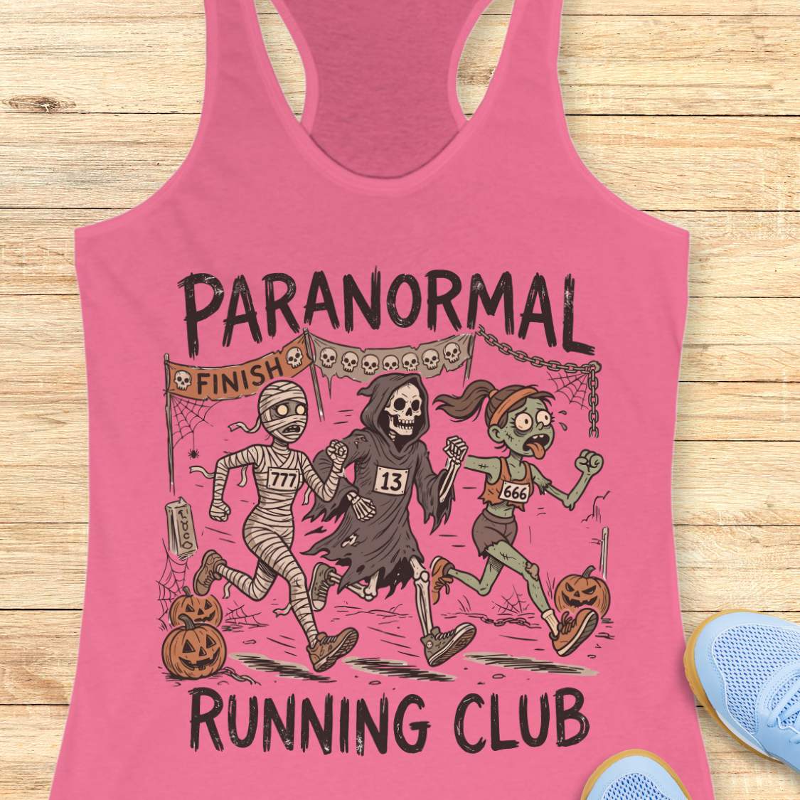 pink Halloween tank top with an image of a running zombie, mummy and reaper,  and text 'Paranormal running club', on a wooden surface