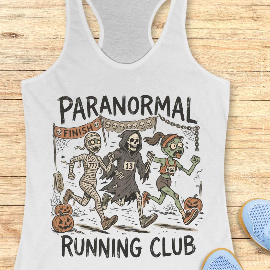 white Halloween tank top with an image of a running zombie, mummy and reaper,  and text 'Paranormal running club', on a wooden surface