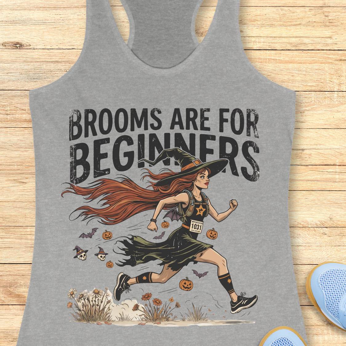 A heather gray racerback tank top featuring a Halloween graphic with the text 'BROOMS ARE FOR BEGINNERS' and an illustration of a witch running with brooms. The tank top is displayed flat on a light wood background with blue shoes partially visible at the bottom right.