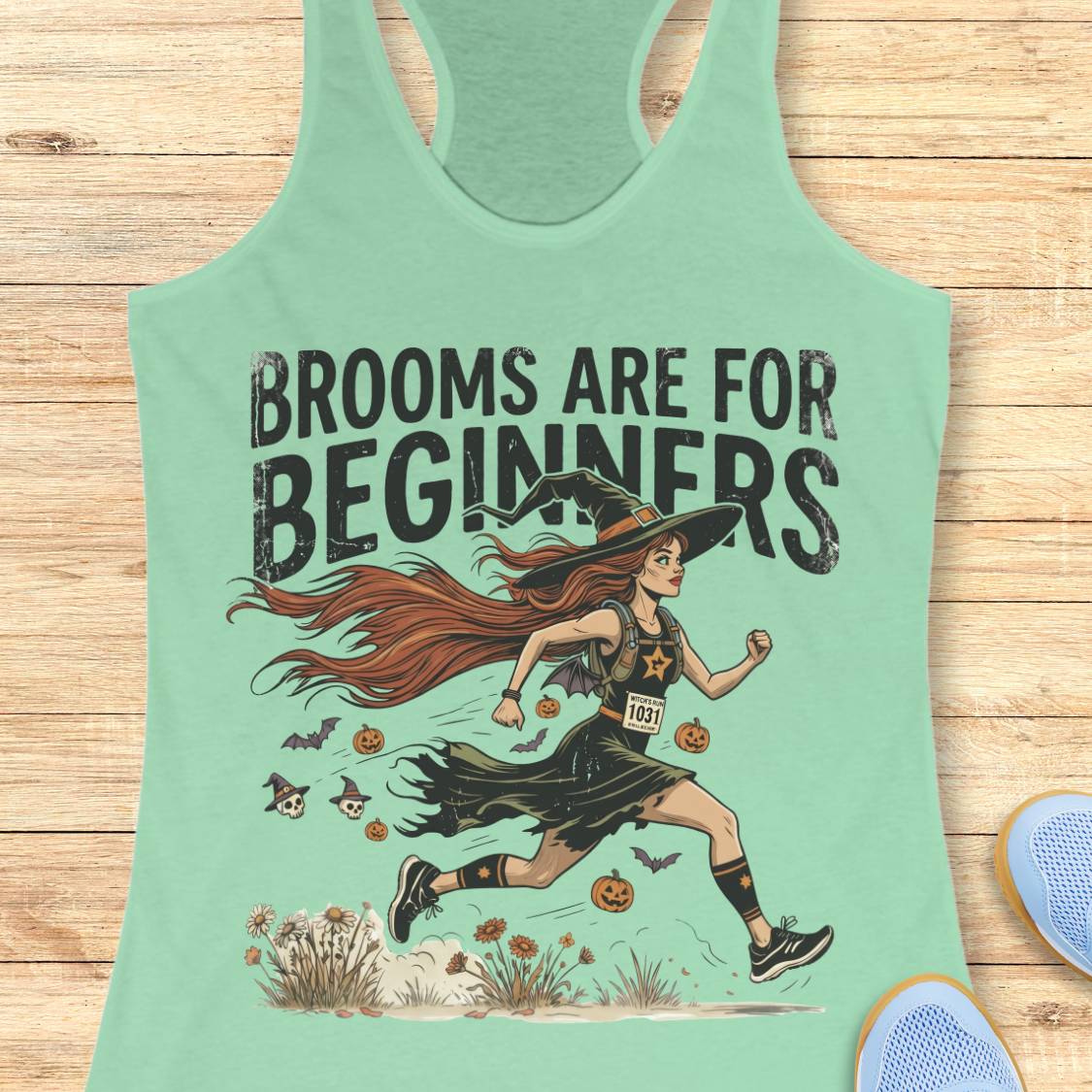 A mint green racerback tank top with a graphic of a witch running with the text 'BROOMS ARE FOR BEGINNERS' on a light wood background.
