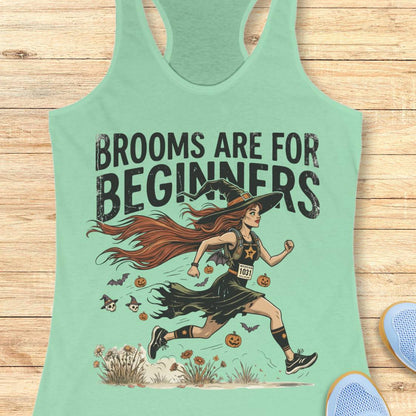 A mint green racerback tank top with a graphic of a witch running with the text 'BROOMS ARE FOR BEGINNERS' on a light wood background.
