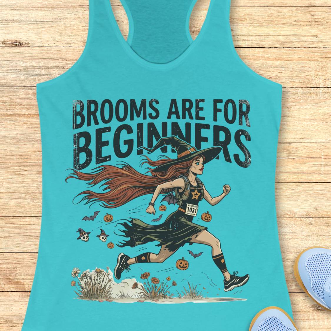 A turquoise racerback tank top laid flat on a light wood surface, featuring a Halloween graphic of a witch running with the text 'BROOMS ARE FOR BEGINNERS.' A blue shoe is partially visible at the lower right edge.
