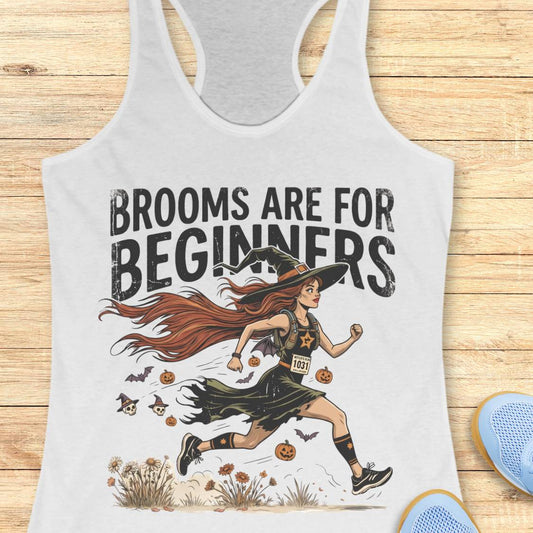 A white racerback tank top laid flat on a light wood plank background. The tank top features a graphic of a woman running as a witch with the text 'BROOMS ARE FOR BEGINNERS' in distressed black lettering.