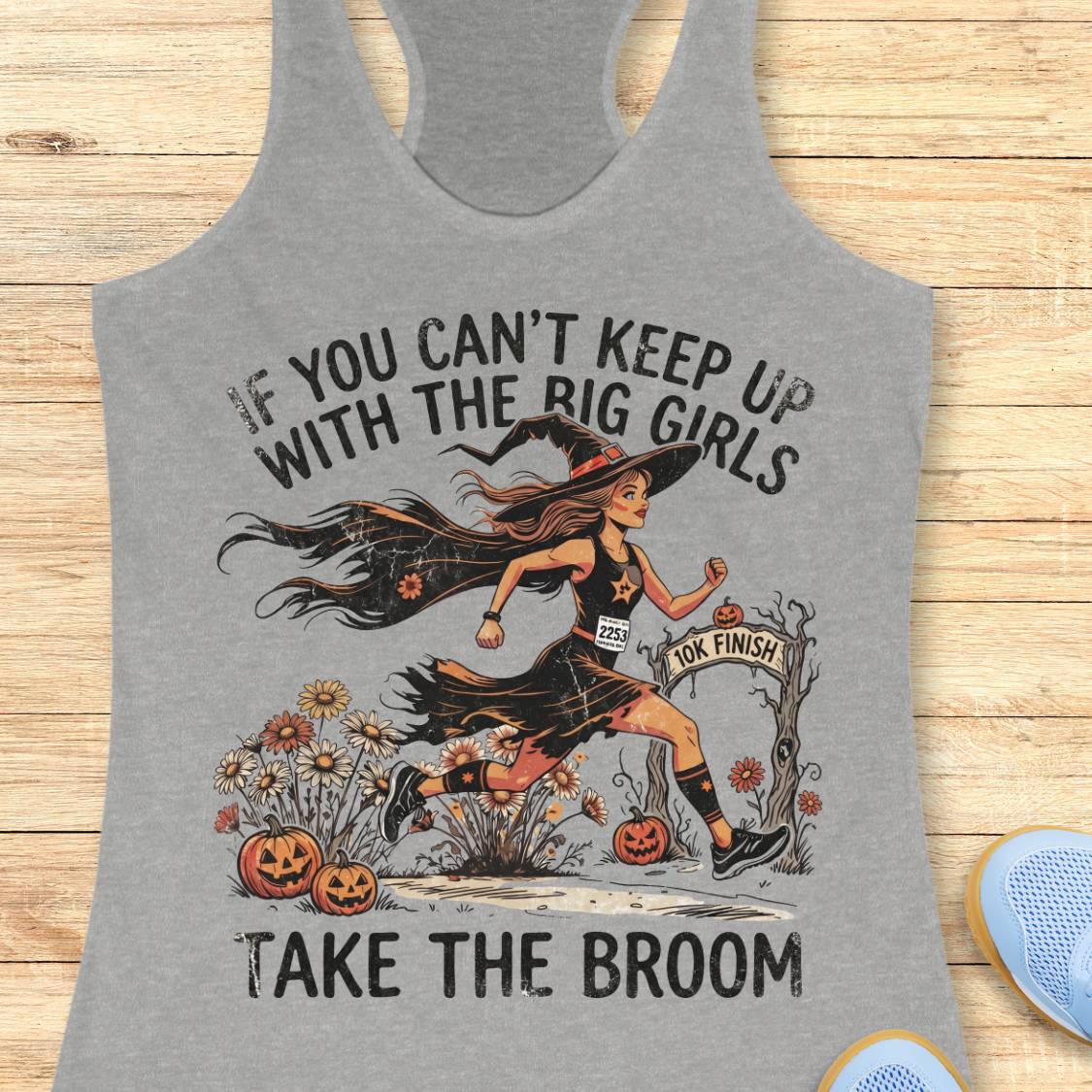 A heather gray racerback tank top laid flat on a light wood surface, featuring a Halloween graphic of a witch running with pumpkins and the text 'IF YOU CAN'T KEEP UP WITH THE BIG GIRLS TAKE THE BROOM'.