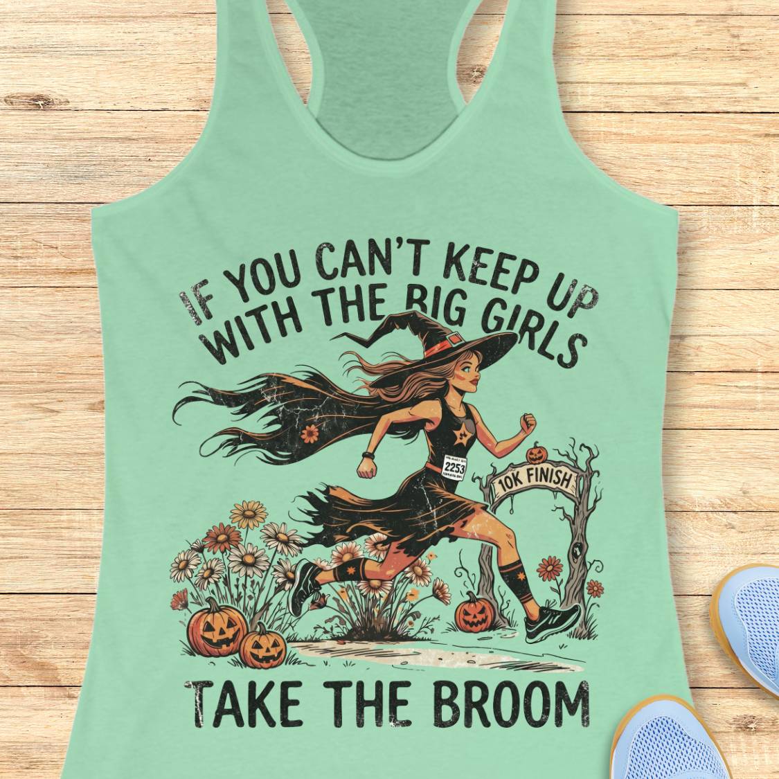 A mint green racerback tank top laid flat on a light wood background, featuring a Halloween graphic of a witch running with a broom and the text 'IF YOU CAN'T KEEP UP WITH THE BIG GIRLS TAKE THE BROOM'. Partial blue shoes are visible at the bottom right edge.