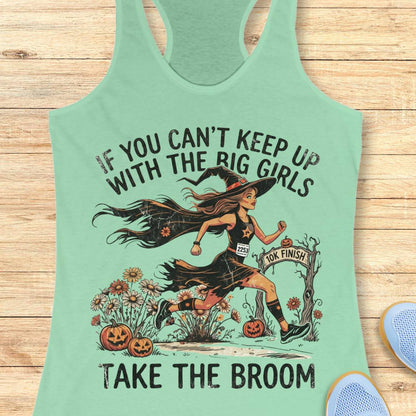 A mint green racerback tank top laid flat on a light wood background, featuring a Halloween graphic of a witch running with a broom and the text 'IF YOU CAN'T KEEP UP WITH THE BIG GIRLS TAKE THE BROOM'. Partial blue shoes are visible at the bottom right edge.