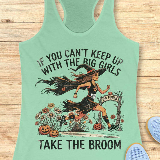 A mint green racerback tank top laid flat on a light wood background, featuring a Halloween graphic of a witch running with a broom and the text 'IF YOU CAN'T KEEP UP WITH THE BIG GIRLS TAKE THE BROOM'. Partial blue shoes are visible at the bottom right edge.
