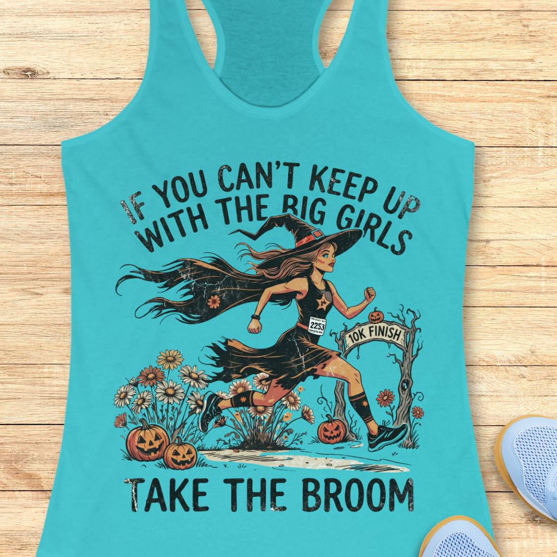 A turquoise racerback tank top laid flat on a light wood surface, featuring a Halloween graphic of a witch running with the text 'IF YOU CAN'T KEEP UP WITH THE BIG GIRLS TAKE THE BROOM.' A small blue sneaker is partially visible at the lower right edge.