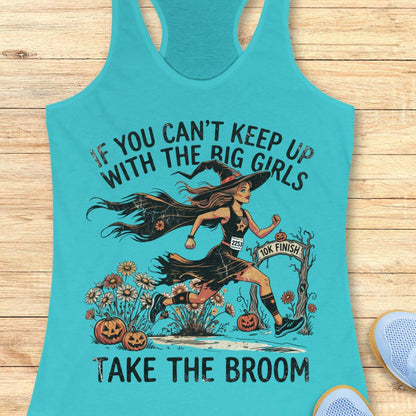 A turquoise racerback tank top laid flat on a light wood surface, featuring a Halloween graphic of a witch running with the text 'IF YOU CAN'T KEEP UP WITH THE BIG GIRLS TAKE THE BROOM.' A small blue sneaker is partially visible at the lower right edge.