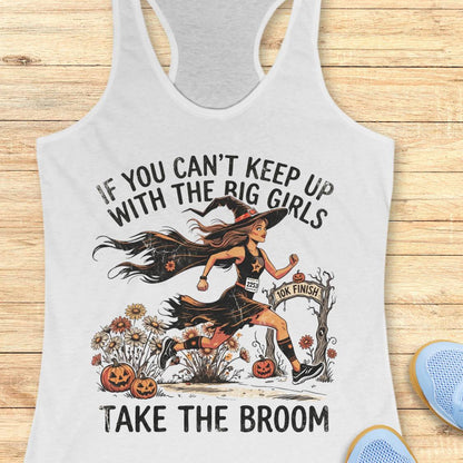 A white racerback tank top laid flat on a light wood background, featuring a Halloween graphic of a witch running with pumpkins and the text 'IF YOU CAN'T KEEP UP WITH THE BIG GIRLS TAKE THE BROOM'. Part of a blue shoe is visible at the bottom right.