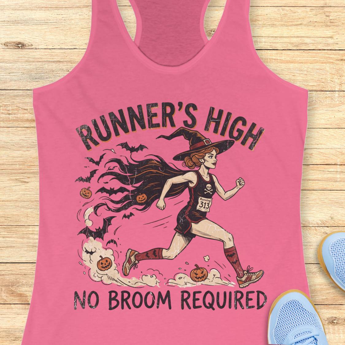A pink racerback tank top laid flat on a light wood surface, featuring a Halloween-themed graphic of a woman running as a witch with the text 'RUNNER'S HIGH' and 'NO BROOM REQUIRED.' Partial blue shoes are visible at the edges as styling props.