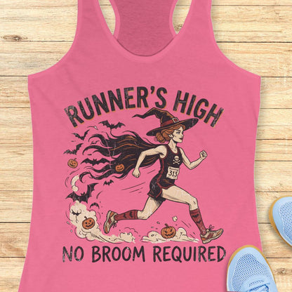 A pink racerback tank top laid flat on a light wood surface, featuring a Halloween-themed graphic of a woman running as a witch with the text 'RUNNER'S HIGH' and 'NO BROOM REQUIRED.' Partial blue shoes are visible at the edges as styling props.