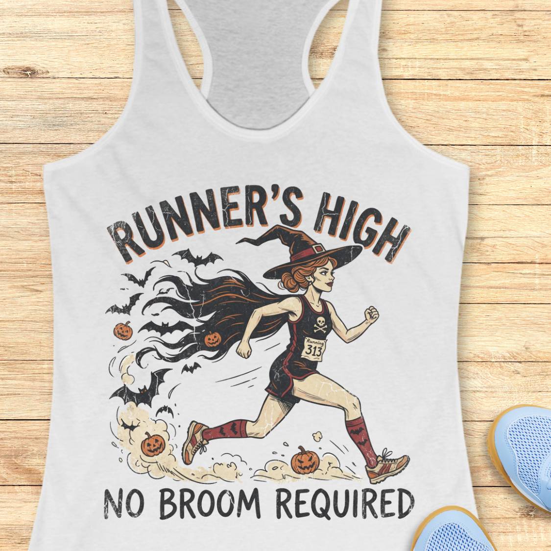 A white racerback tank top laid flat on a light wood surface, featuring a Halloween-themed graphic of a witch running with bats, pumpkins, and the text 'RUNNER'S HIGH NO BROOM REQUIRED.' Partial blue shoes are visible at the bottom right as styling props.