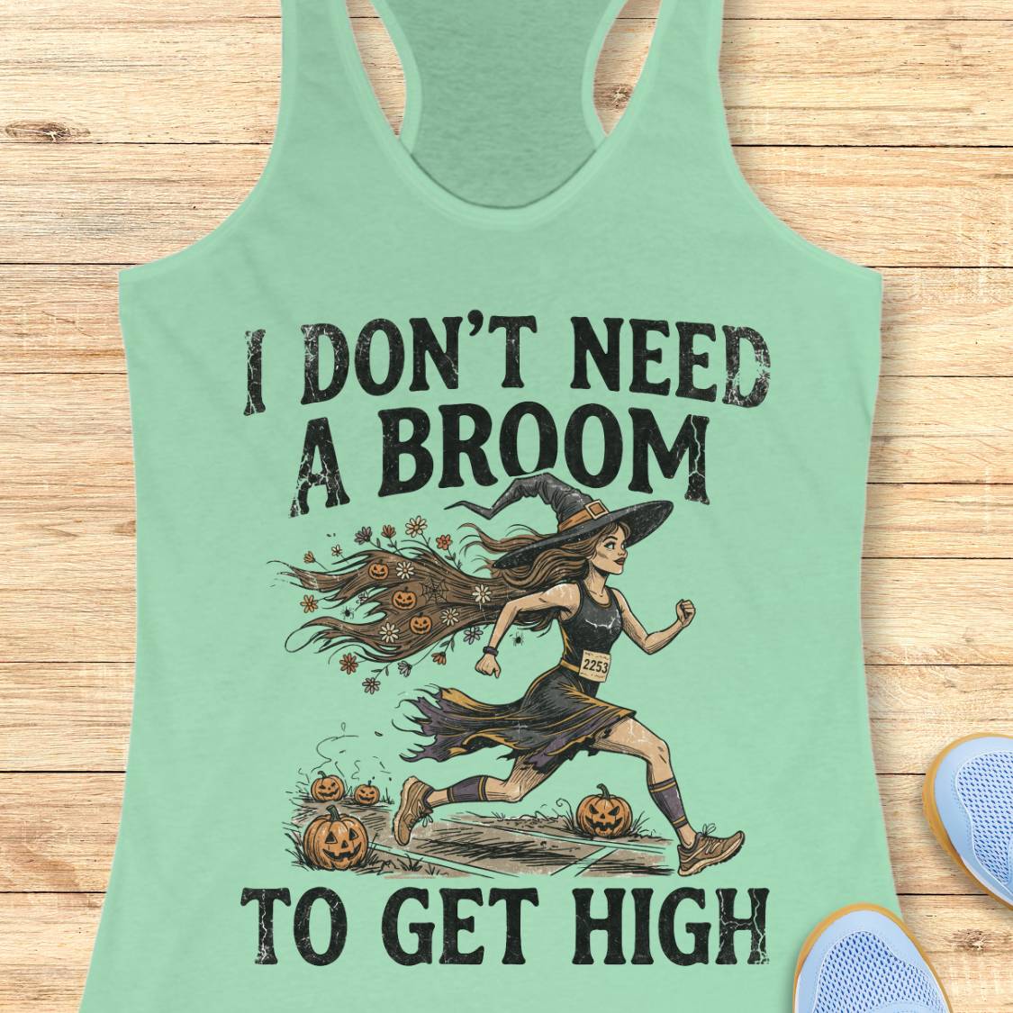 A mint green racerback tank top laid flat on a light wood background, featuring a graphic of a witch running with pumpkins and the text 'I DON'T NEED A BROOM TO GET HIGH'.