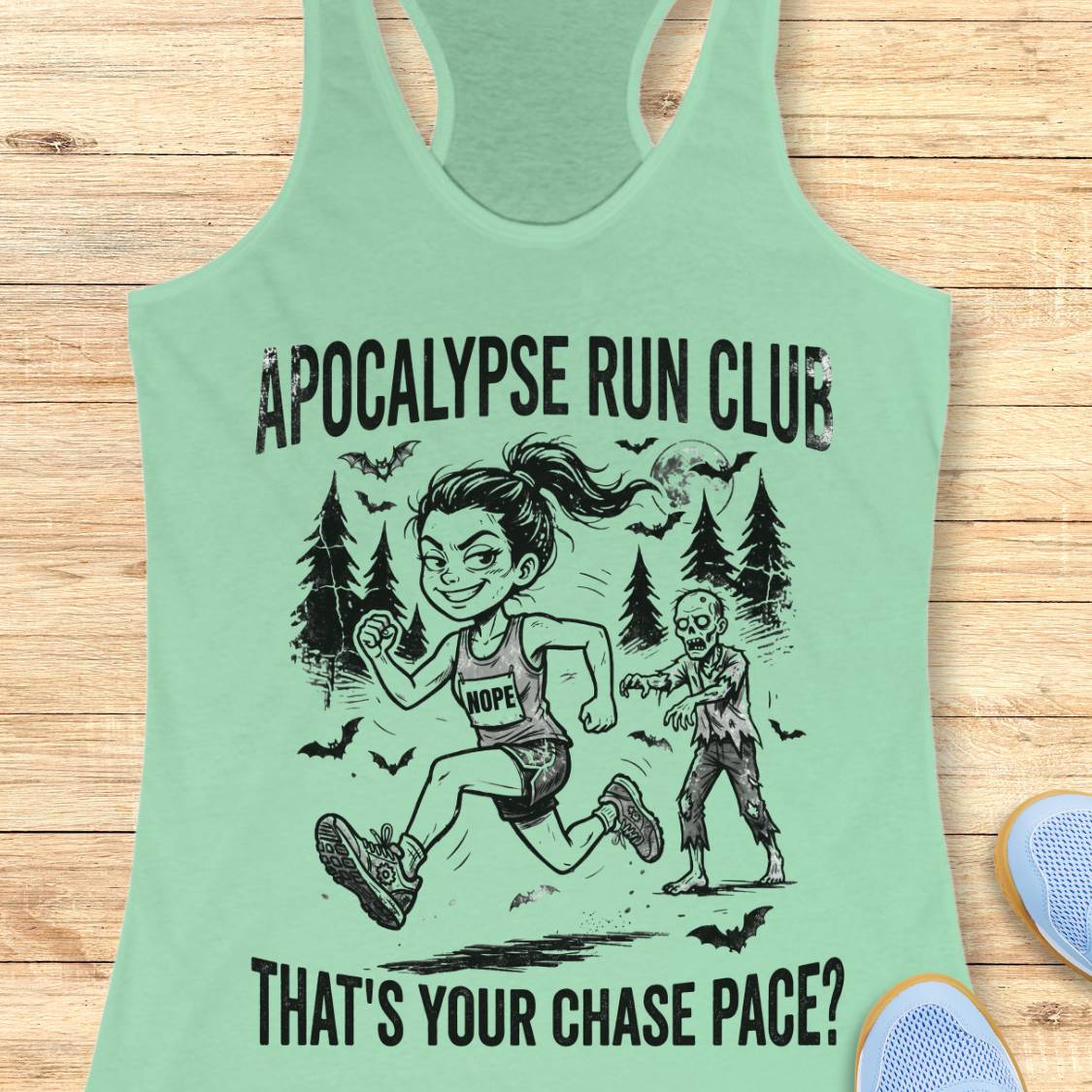 A mint green racerback tank top laid flat on a light wood background, featuring a black graphic of a woman running with a zombie and the text 'APOCALYPSE RUN CLUB' and 'THAT'S YOUR CHASE PACE?'.