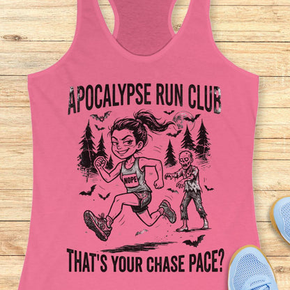 A pink racerback tank top laid flat on a light wood background, featuring a black graphic of a woman running with a zombie and the text 'APOCALYPSE RUN CLUB' and 'THAT'S YOUR CHASE PACE?'. Partial blue shoes are visible at the edges.
