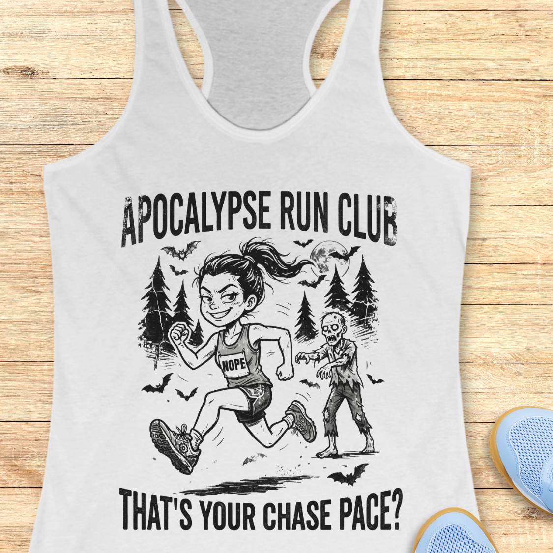 A white racerback tank top laid flat on a light wood surface, featuring a black graphic of a woman running with a zombie and the text 'APOCALYPSE RUN CLUB' and 'THAT'S YOUR CHASE PACE?'. Partial blue shoes are visible at the bottom right.
