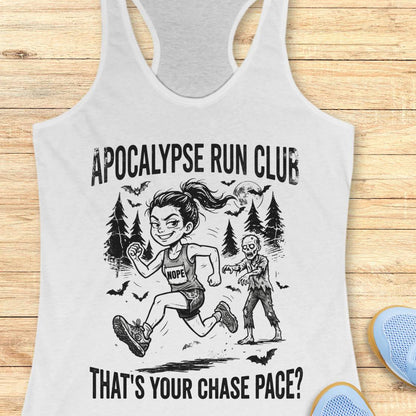 A white racerback tank top laid flat on a light wood surface, featuring a black graphic of a woman running with a zombie and the text 'APOCALYPSE RUN CLUB' and 'THAT'S YOUR CHASE PACE?'. Partial blue shoes are visible at the bottom right.