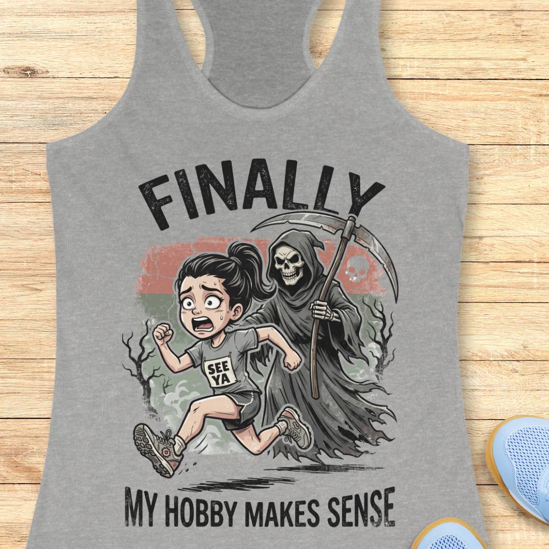 A heather gray racerback tank top laid flat on a light wood surface, featuring a cartoon graphic of a woman running while being chased by a grim reaper, with the text 'FINALLY MY HOBBY MAKES SENSE'.